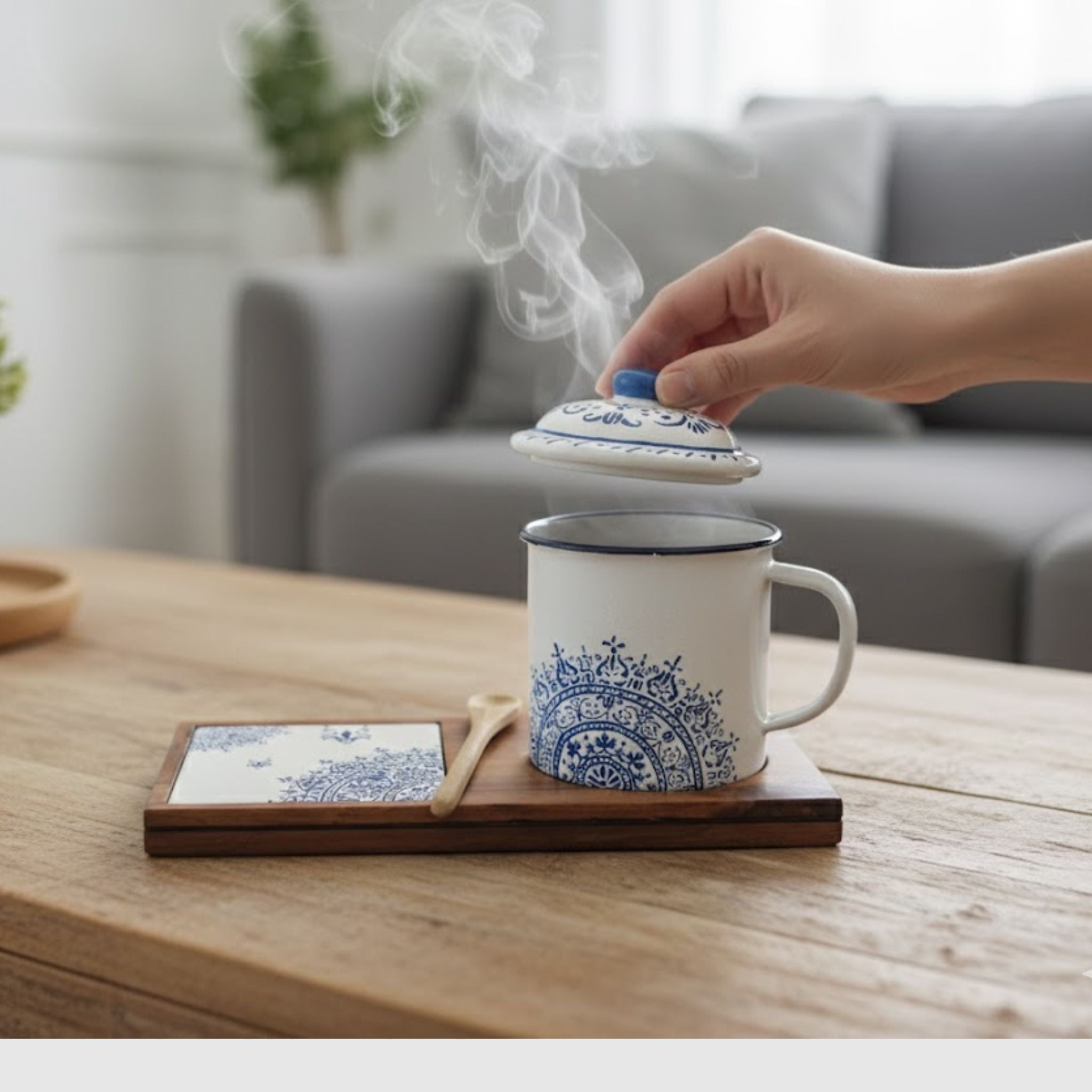 Hand lifting the lid of a steaming Senja Indigo batik mug on its wooden tray with coaster, styled in a living room